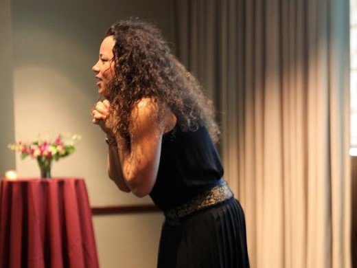 Laura clutching hands and leaning engagingly toward out-of-frame audience in a banquet hall presentation room